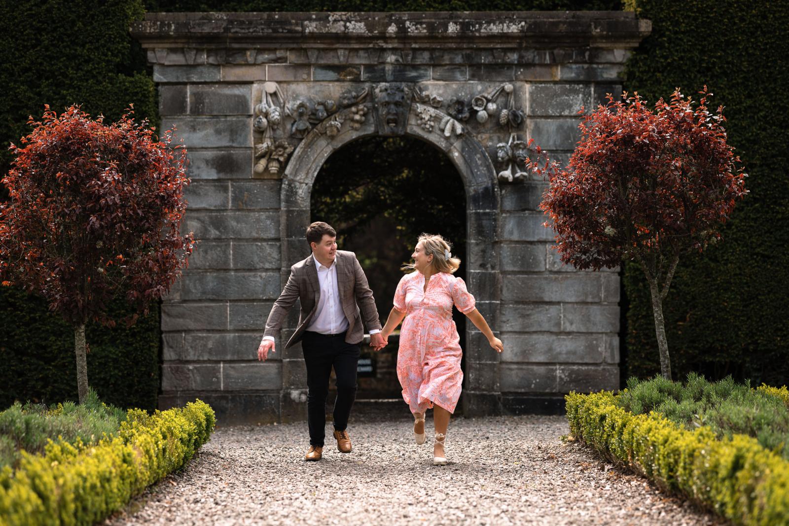 ouple running hand in hand through a historic stone archway in the gardens of Drummond Castle