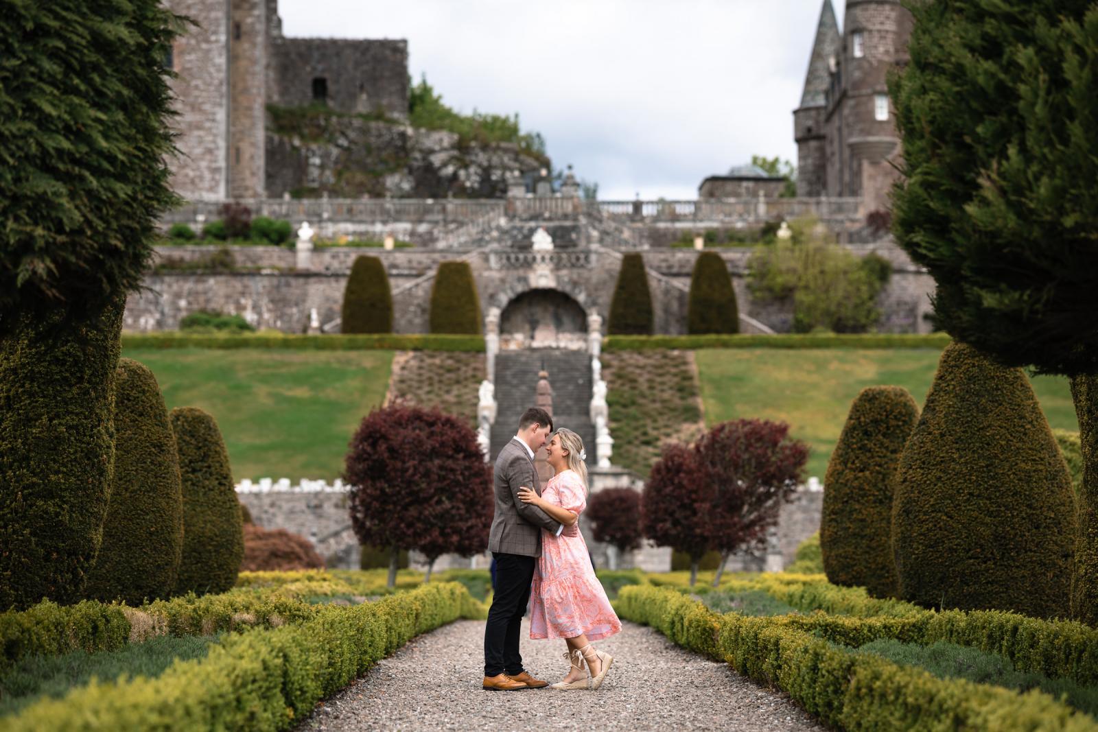 Emma and Hamish embracing on a gravel pathway with the impressive Drummond Castle in the background