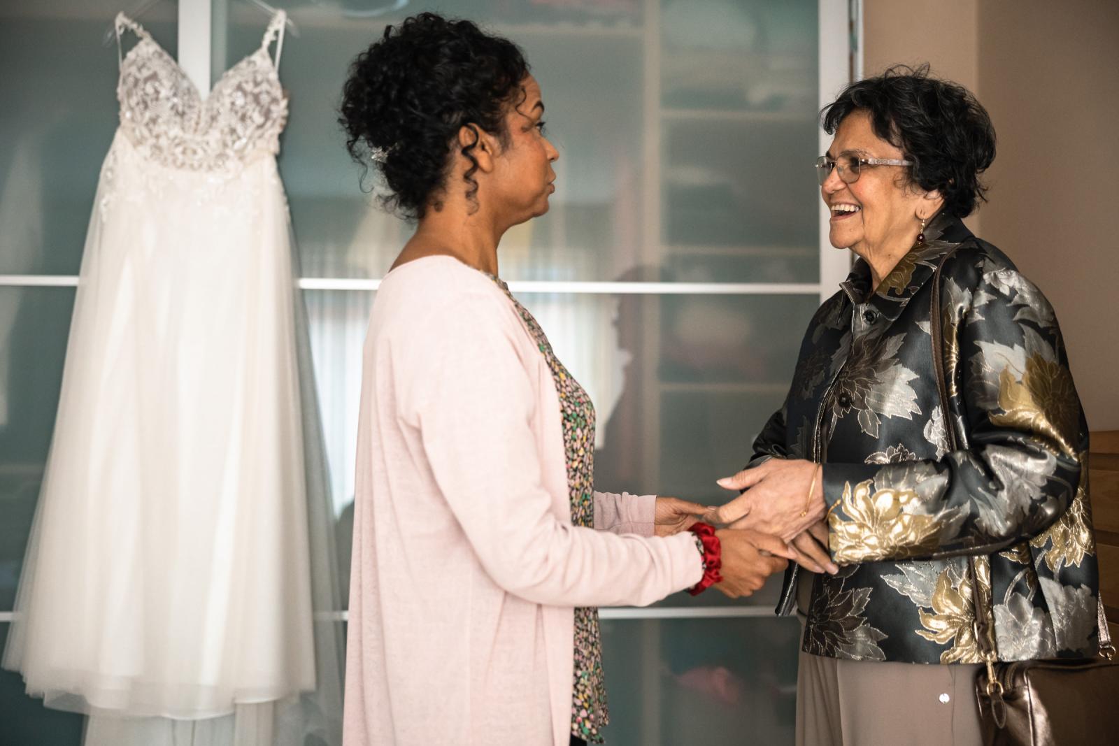 Bruid Farida houdt liefdevol de handen vast van haar moeder terwijl haar trouwjurk op de achtergrond hangt tijdens het aankleedmoment thuis.