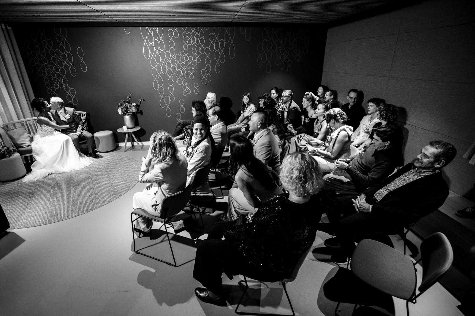 Overzichtsfoto van de trouwzaal met het bruidspaar vooraan en alle gasten aandachtig toekijkend bij de eerste kus.