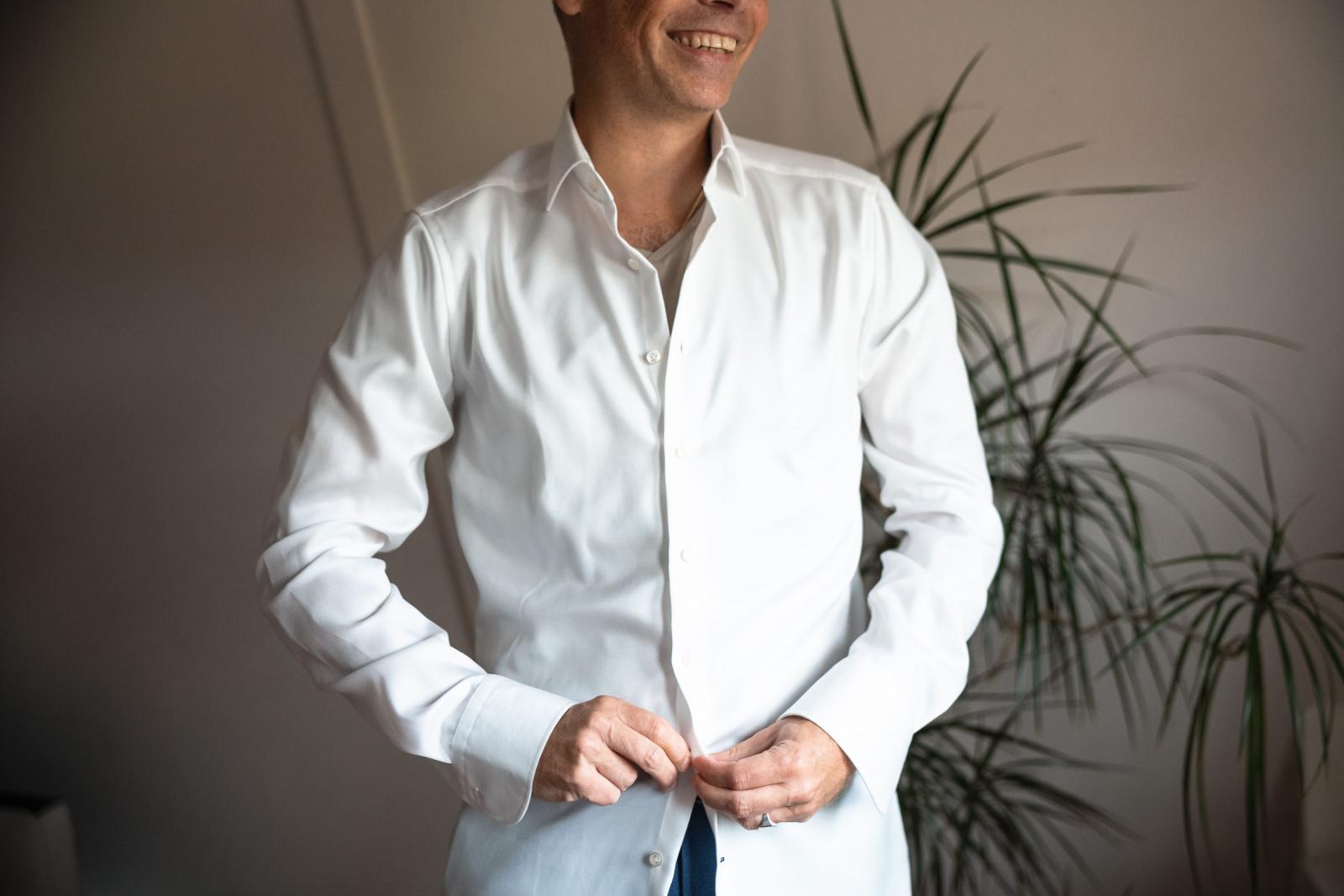 Bruidegom Hans knoopt lachend zijn witte overhemd dicht tijdens de voorbereidingen in de zolderkamer.