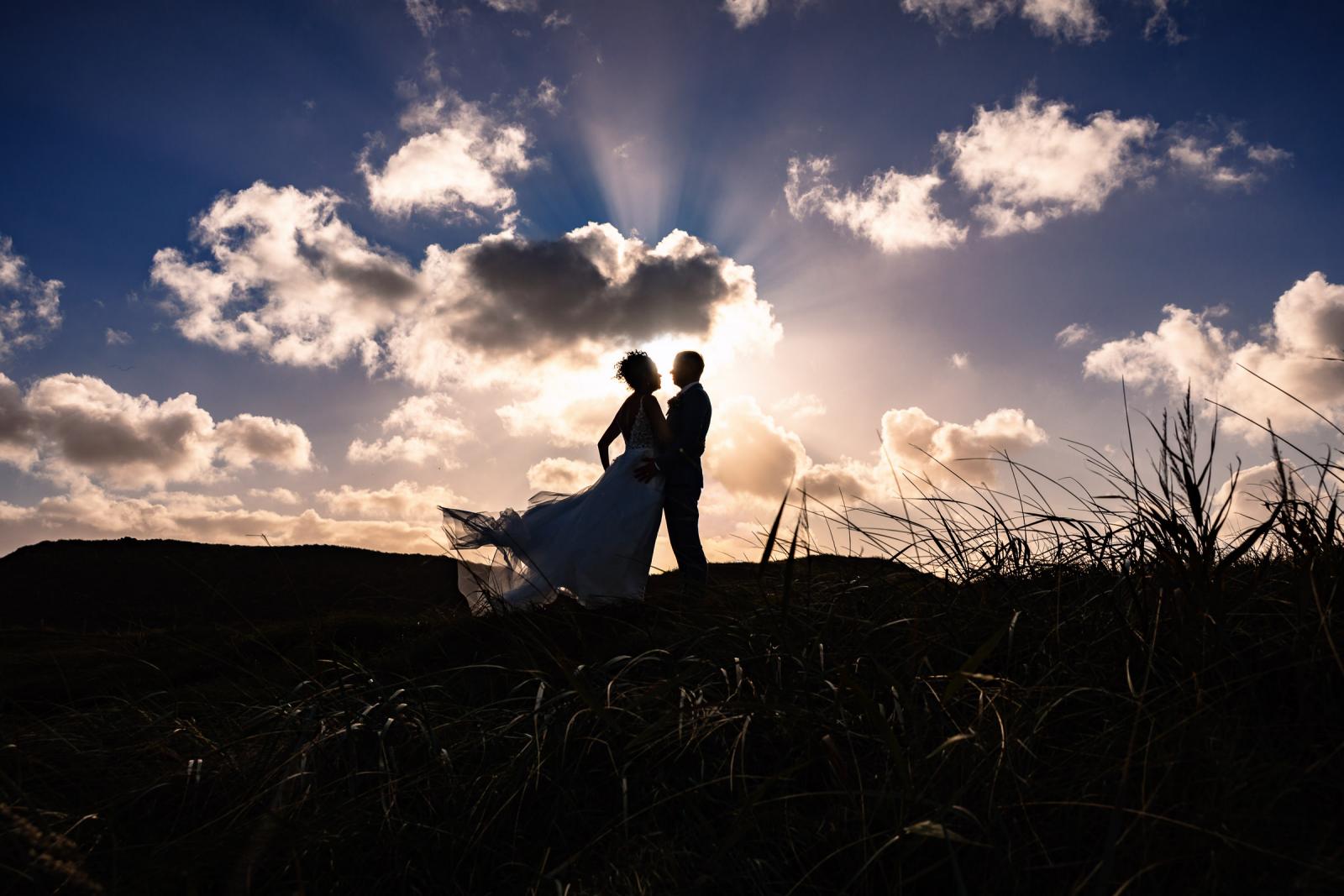 Silhouet van bruidspaar tegen een dramatische lucht tijdens zonsondergang in de duinen.