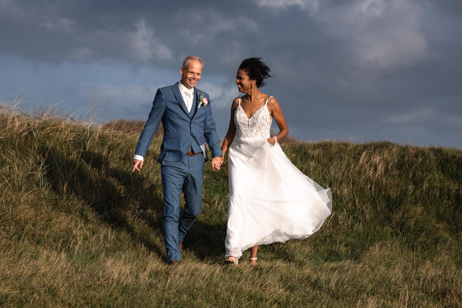 Bruidspaar lacht samen terwijl ze door de duinen lopen onder dreigende wolken.