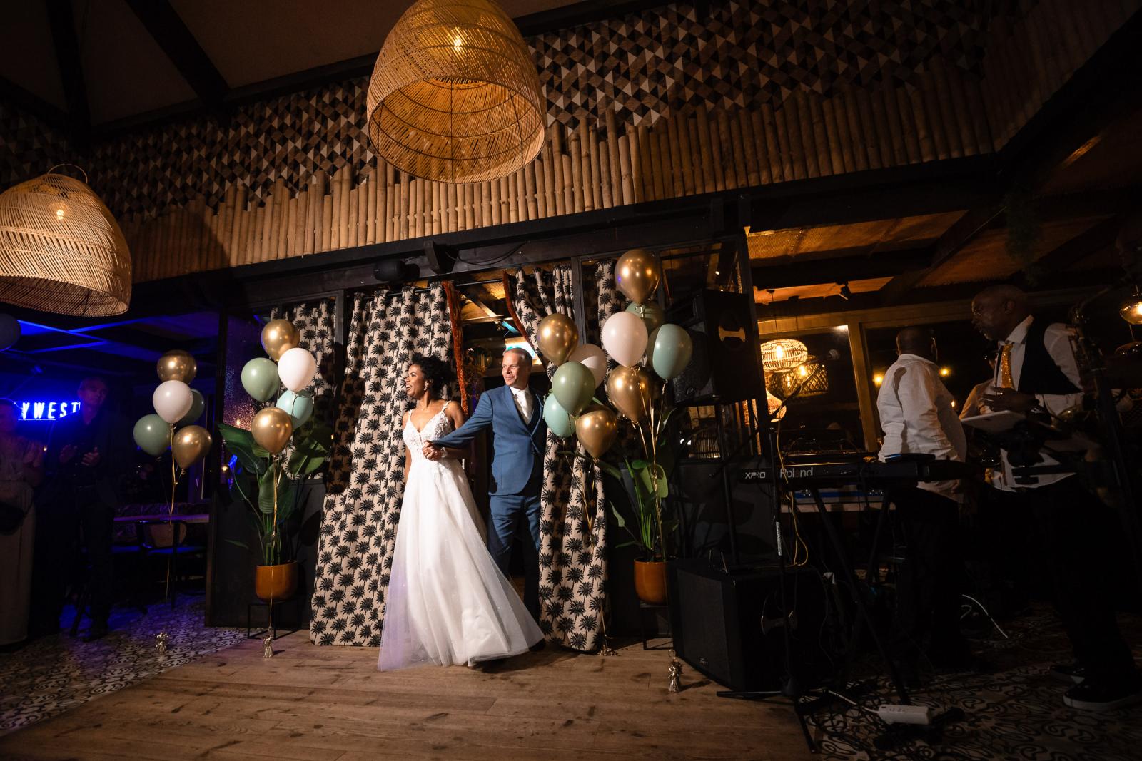 Bruid en bruidegom die de feestzaal binnenkomen tussen ballonnen en warme verlichting, terwijl de band de Zonnebloem op de achtergrond klaarstaat.