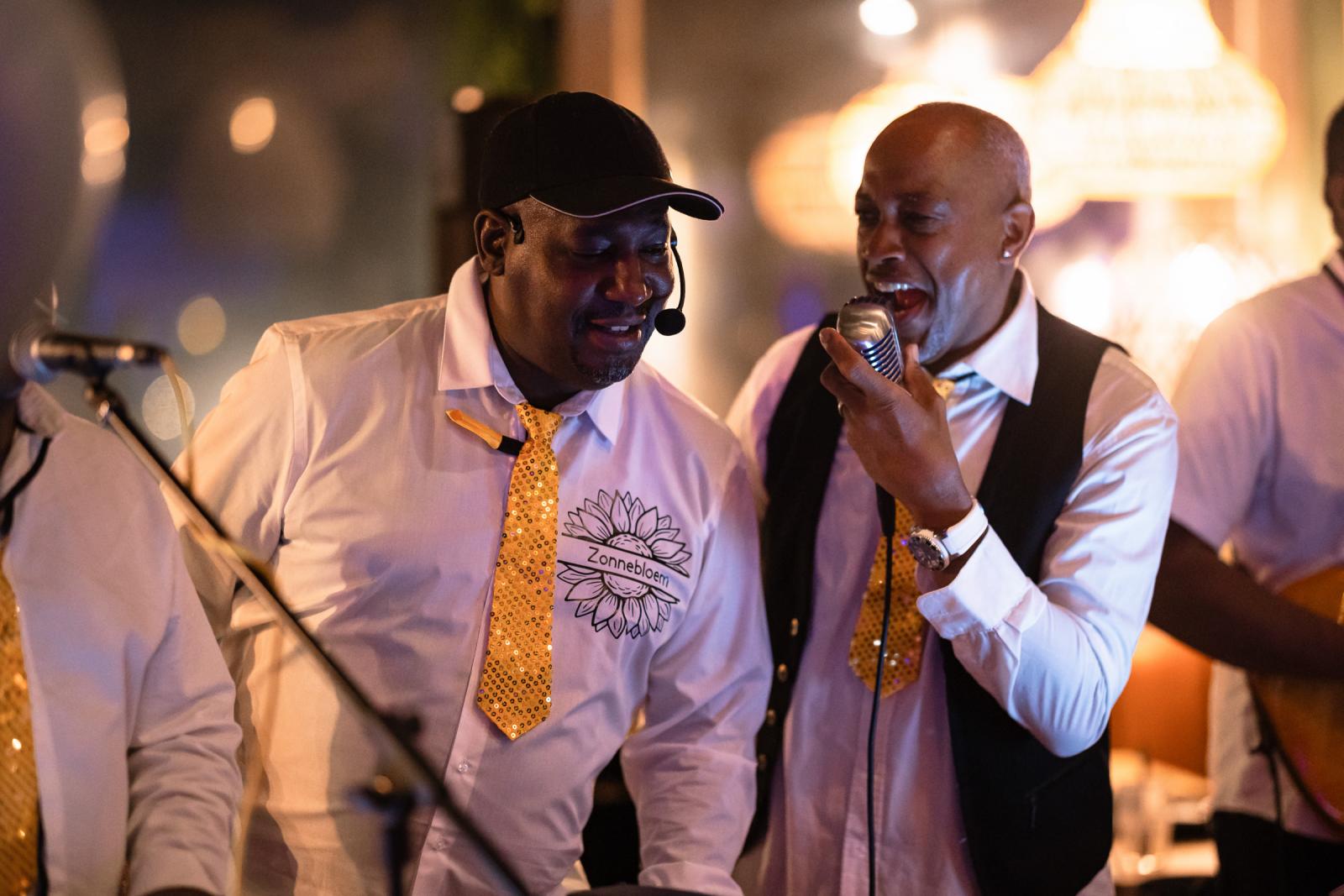Twee zangers van de liveband de Zonnebloem die enthousiast zingen tijdens het trouwfeest, met warme sfeerverlichting.