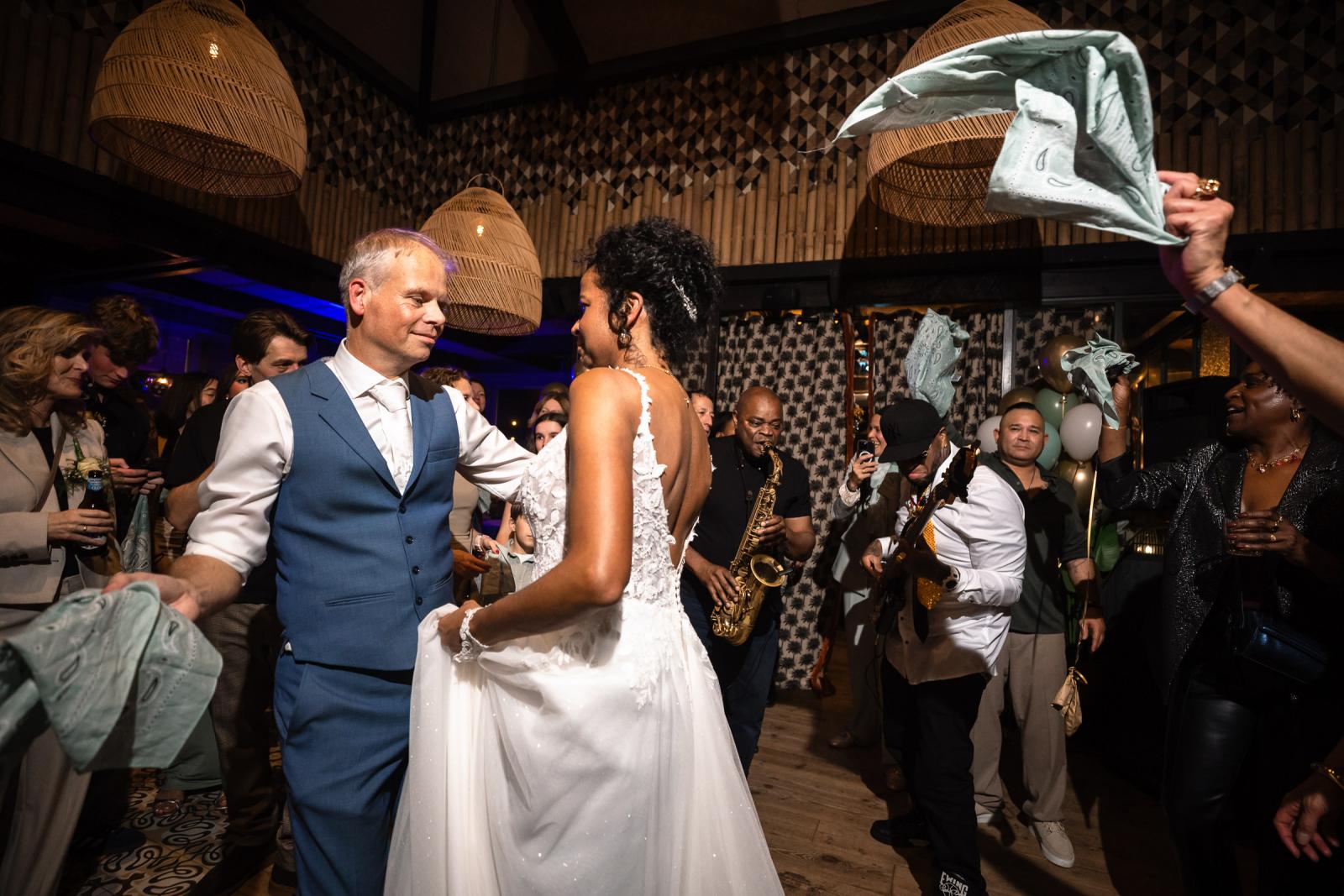 Bruid en bruidegom dansend tussen de gasten terwijl muzikanten live spelen en doeken worden rondgezwaaid.