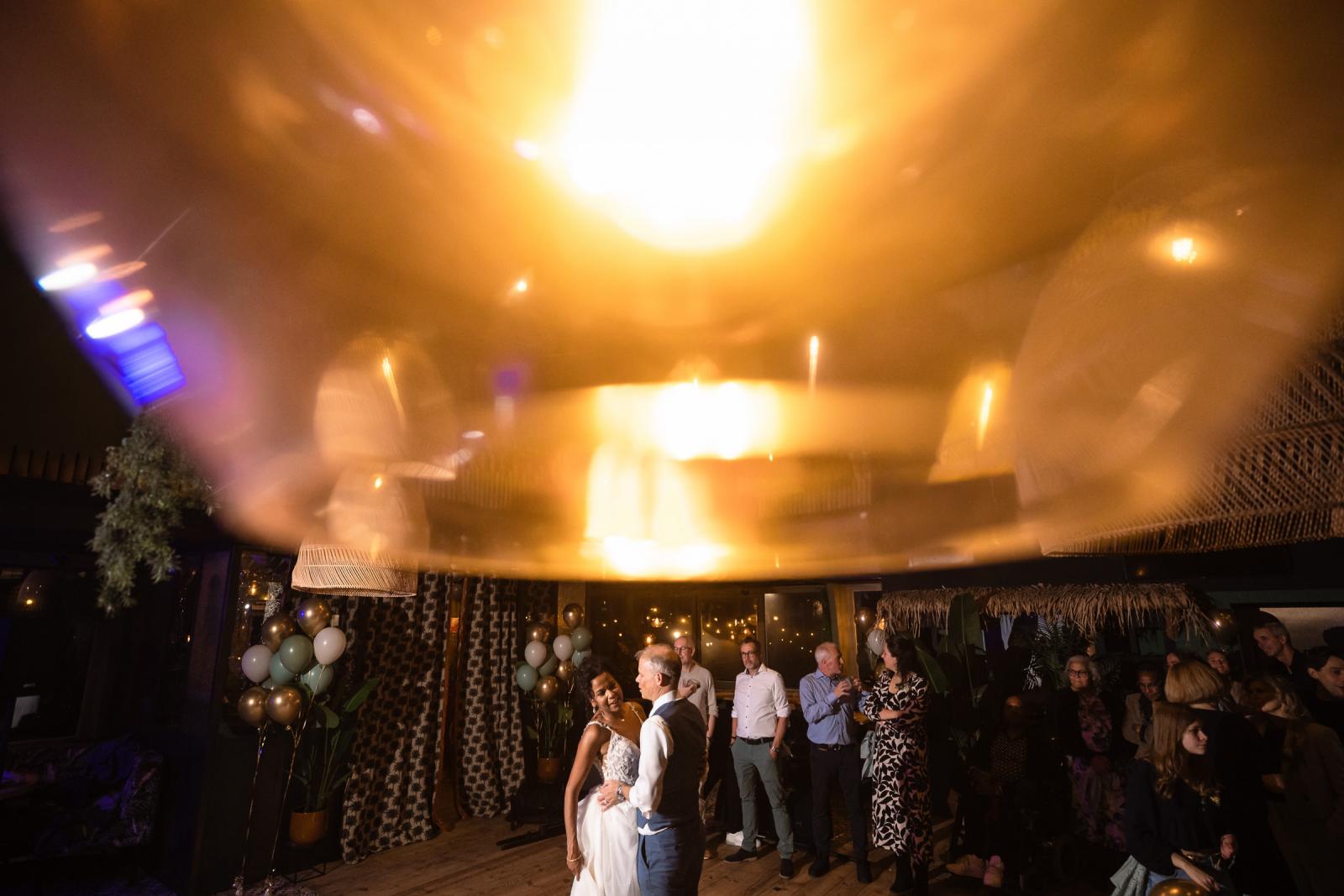 Close-up van warme, gouden lichtreflecties terwijl het bruidspaar danst, omringd door gasten in een sfeervol verlichte feestzaal