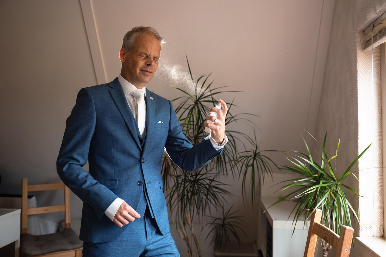 Bruidegom Hans brengt geurig parfum aan op zijn gezicht terwijl hij volledig aangekleed is in zijn blauwe trouwpak.