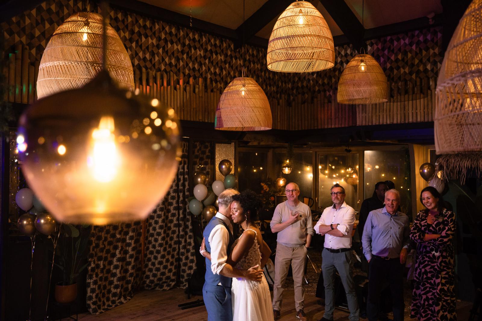 Het bruidspaar danst omringd door toeschouwers in een warm verlichte zaal met grote rotan lampen en feestelijke decoratie
