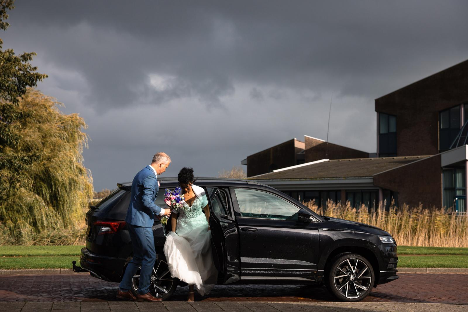 Hans helpt Farida uit de auto stappen bij de trouwlocatie onder een dramatische wolkenlucht.