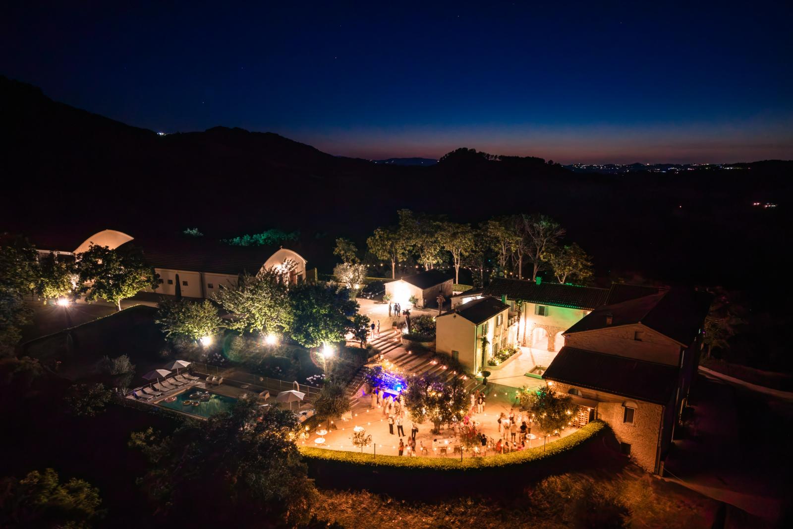 Night drone view of illuminated Le Stonghe venue courtyard decorated with string lights in Le Marche