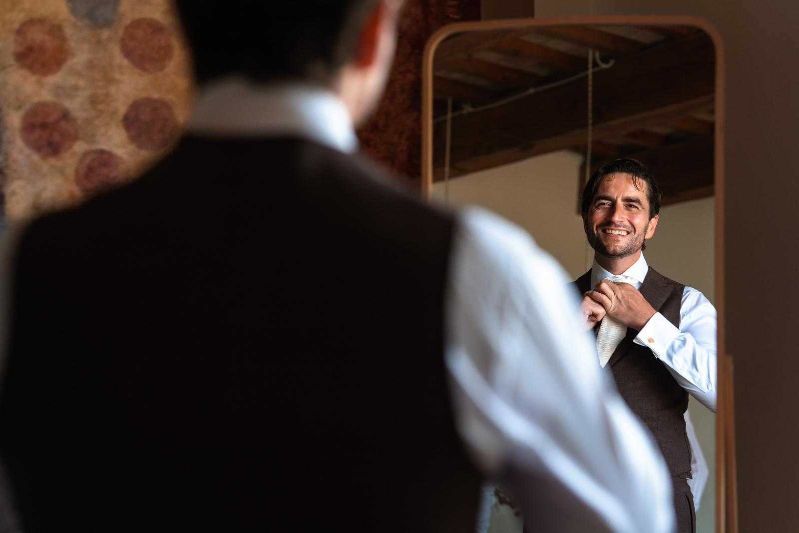 Groom Niels adjusting his tie in a mirror while wearing a stylish brown three-piece wedding suit