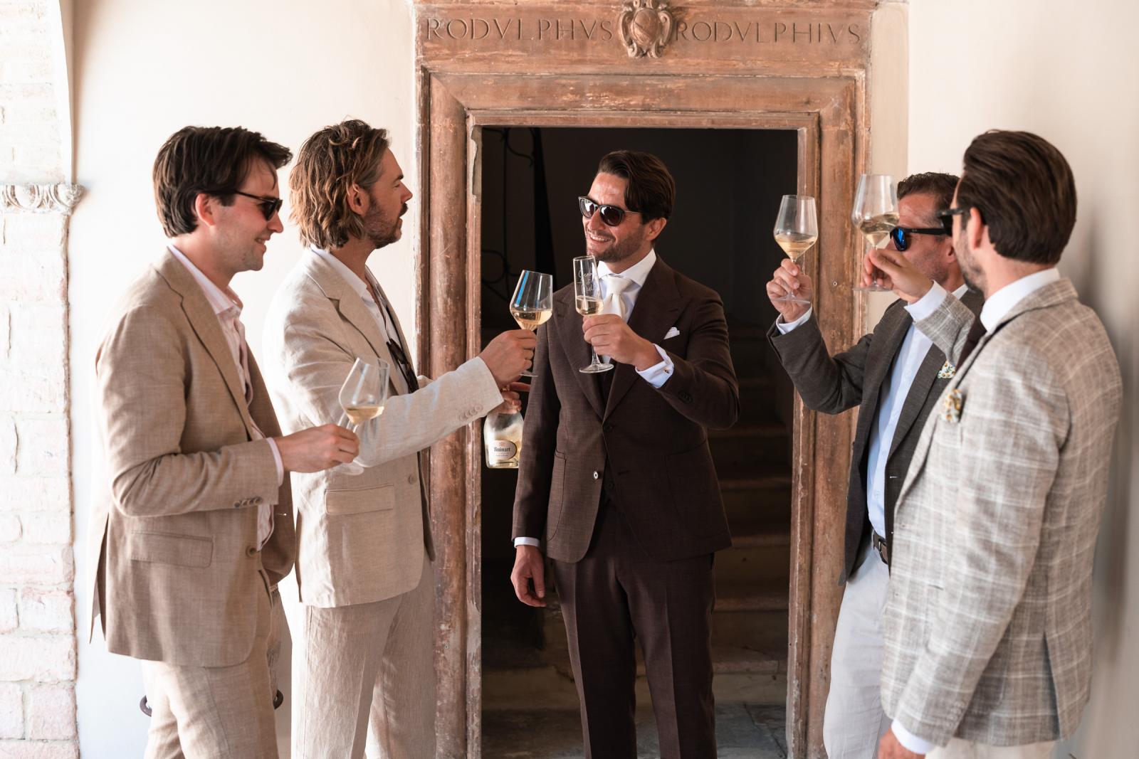 Groom Niels and groomsmen sharing a prosecco toast in front of historic stone archway at Le Stonghe