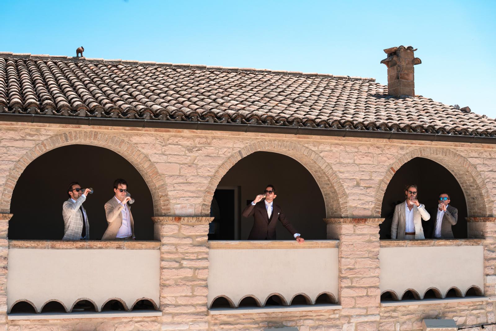 Groom Niels and groomsmen enjoying drinks along the arched stone terrace overlooking Le Marche hills