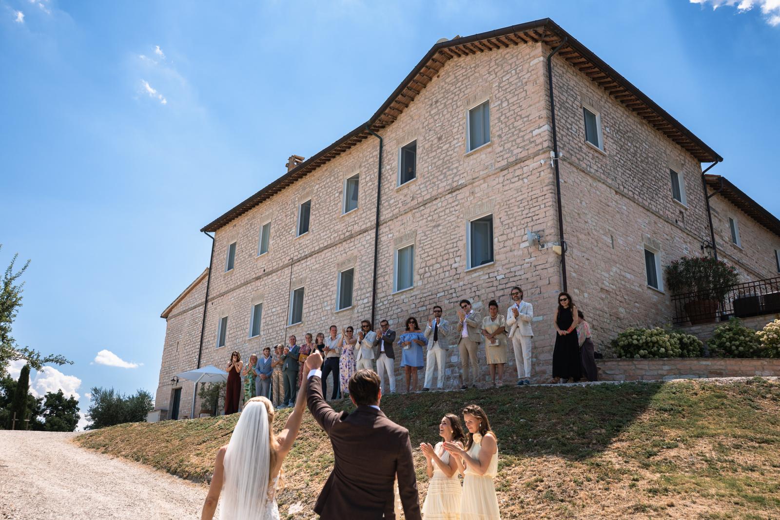 Bride and groom raising hands in celebration while guests cheer from historic stone building terrace