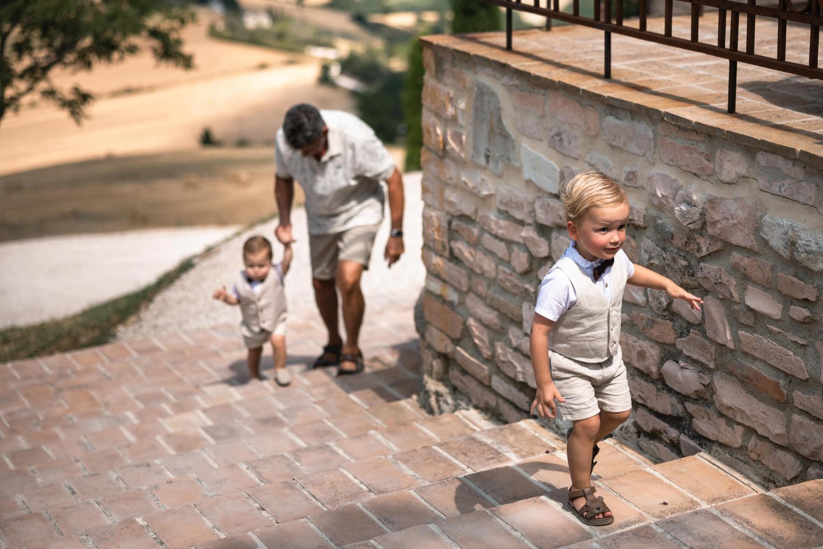 Young page boy walking up brick outdoor steps while grandfather assists another child