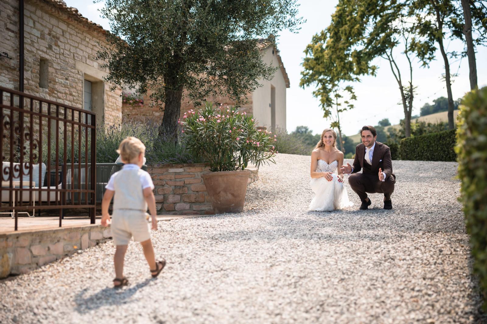 Bride and groom crouching down on gravel path welcoming young page boy running toward them