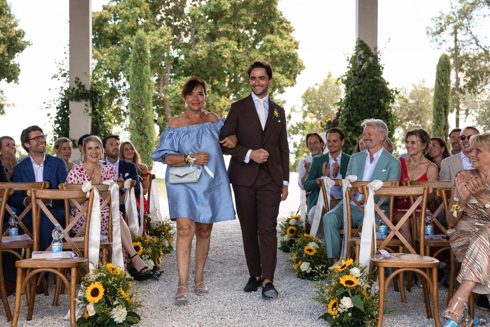 Groom Niels walking down the aisle escorted by his mother wearing a blue dress