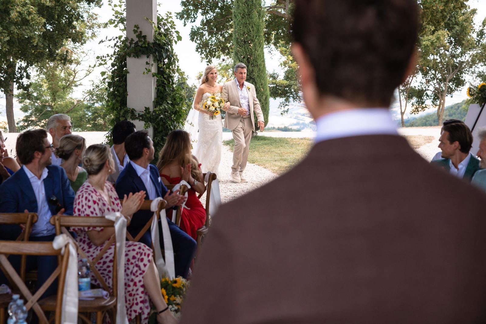 Bride Gemmy walking down gravel aisle with father seen from behind groom Niels