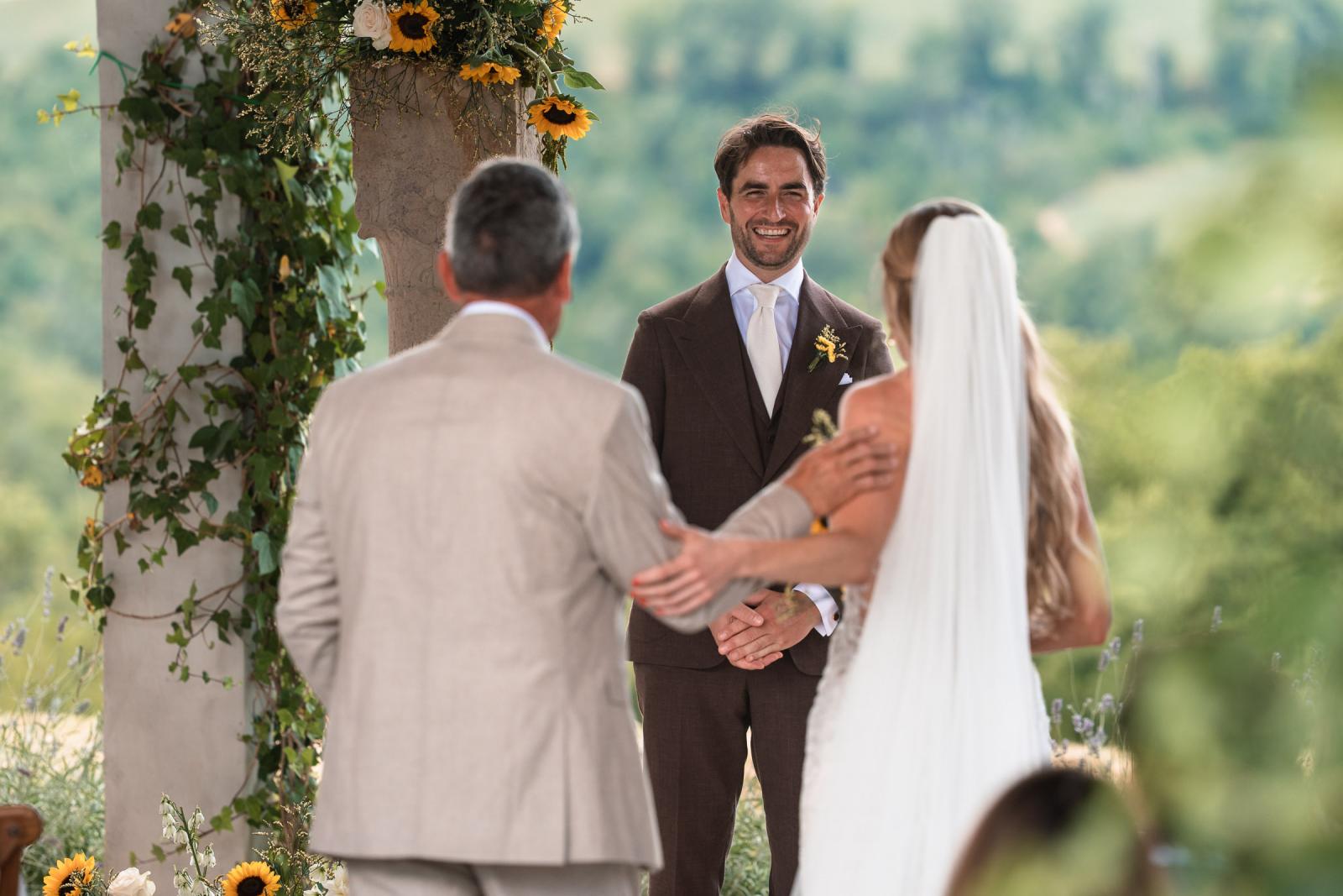 Father handing off bride Gemmy to groom Niels under floral sunflower arch