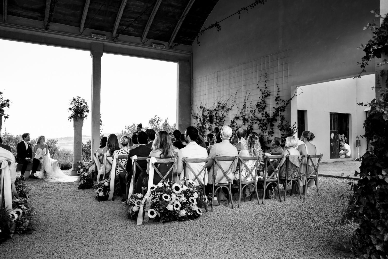 Black and white wide shot of wedding guests seated under covered terrace during ceremony