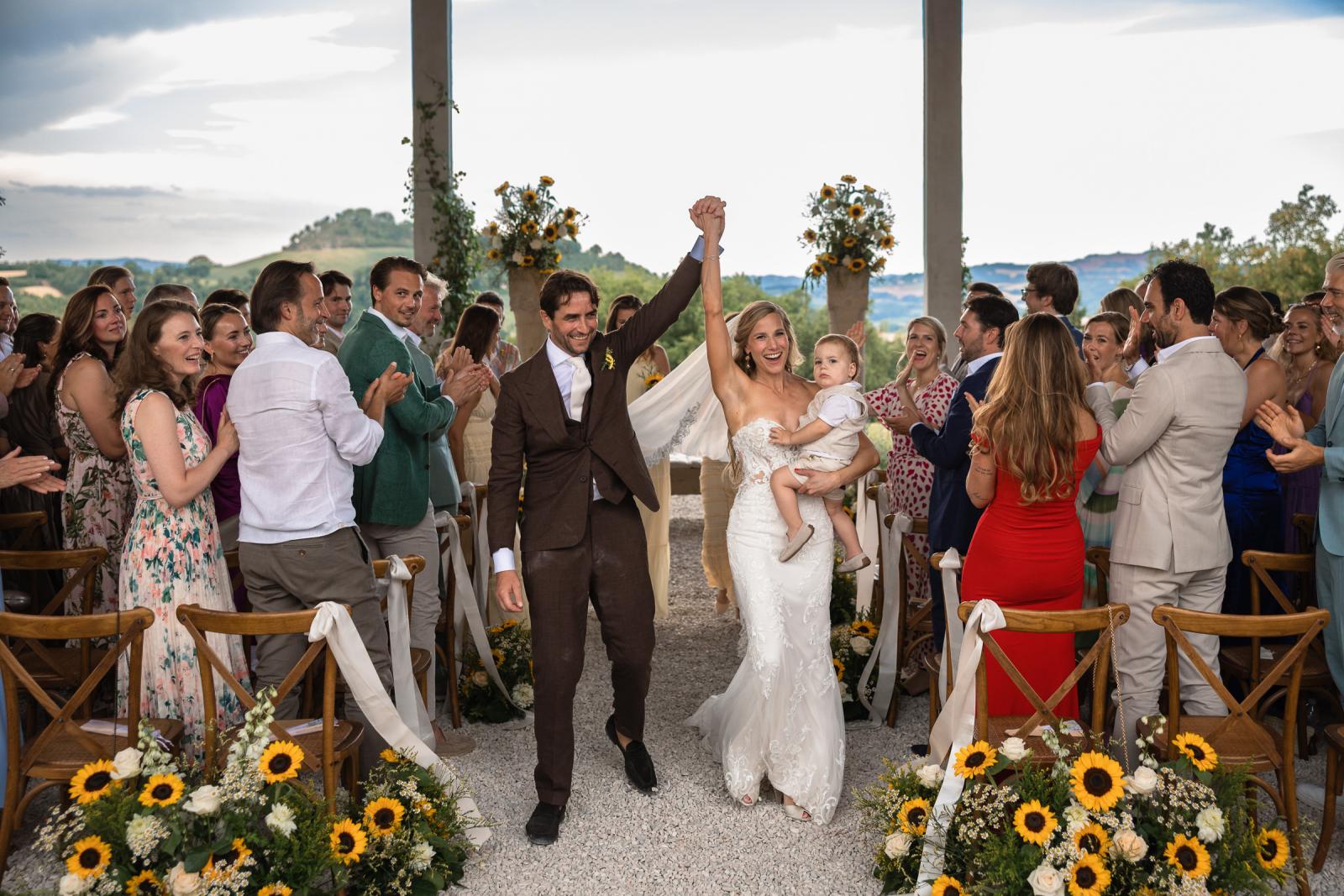 Newlyweds Gemmy and Niels walking down aisle carrying toddler as wedding guests applaud