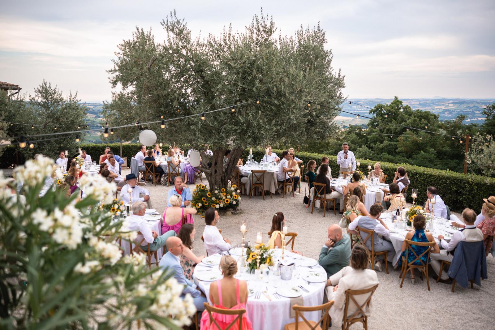 Outdoor wedding dinner setup with round tables under olive trees overlooking Italian landscape