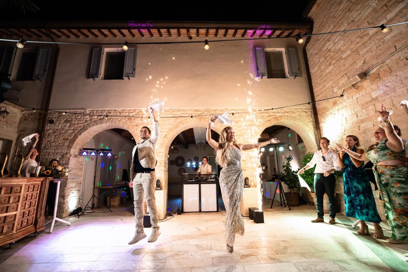 Groom Niels jumping in air and bride Gemmy dancing in courtyard with cold sparkler fountains behind