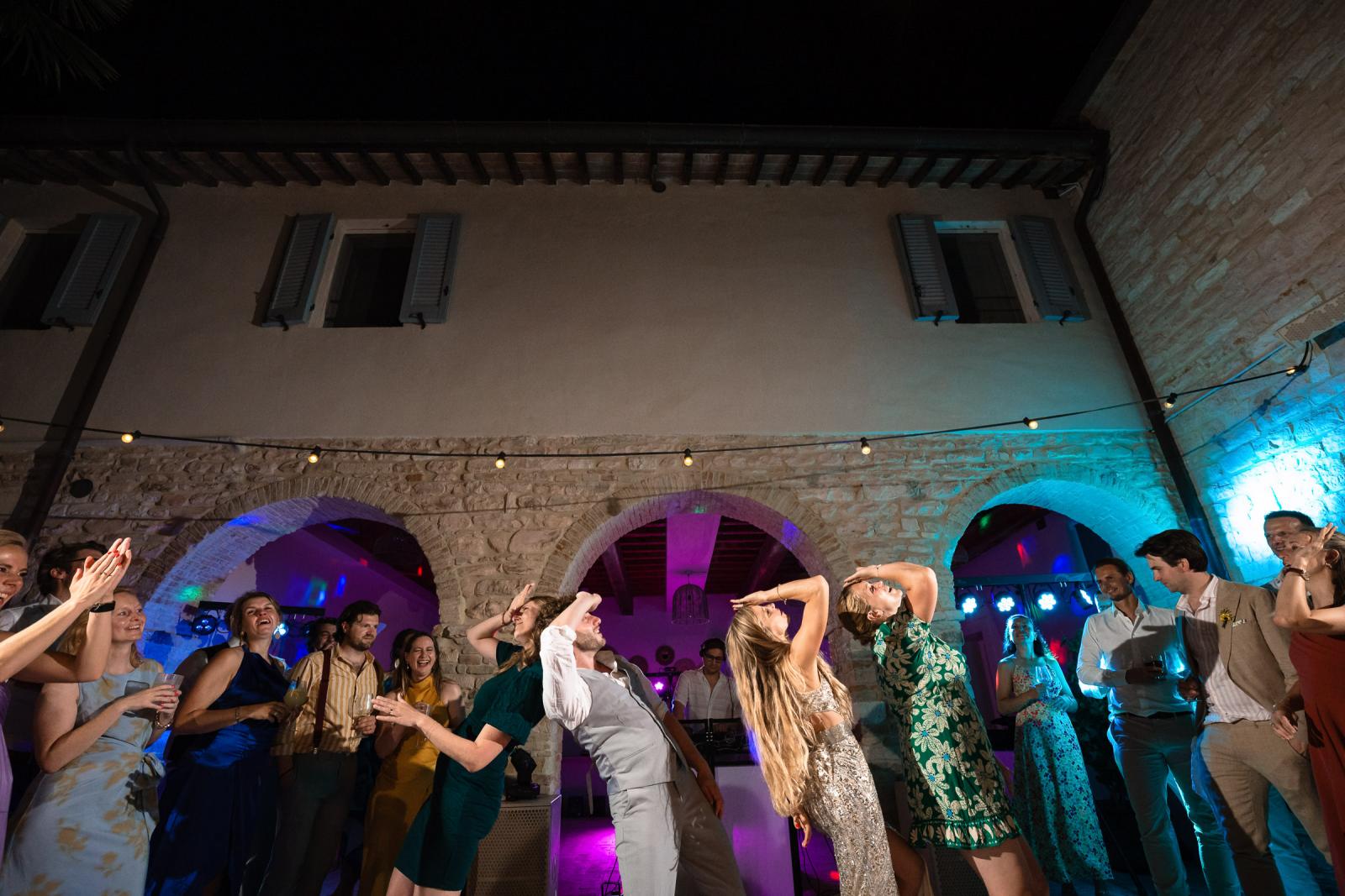 Bride Gemmy and guests leaning backward in synchronized dance line under courtyard arches