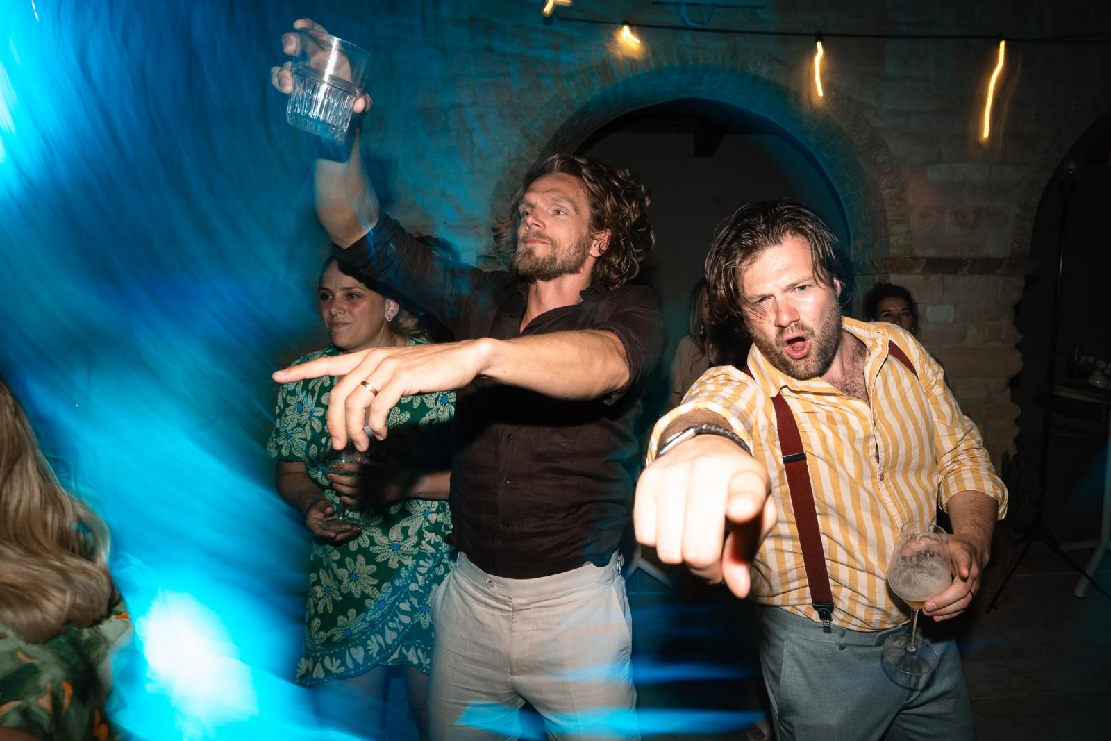 Motion blur capture of male guests dancing energetically with drinks on courtyard dance floor