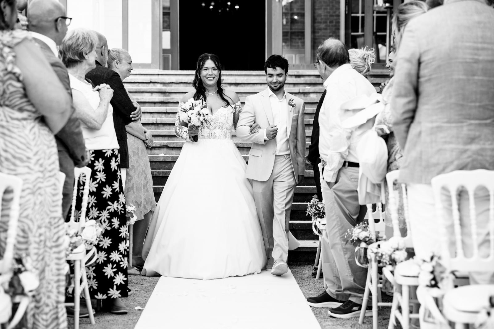 Bruid Iris loopt arm in arm met haar broer over de witte loper naar voren tijdens de buitenceremonie bij Kasteel de Wittenburg.