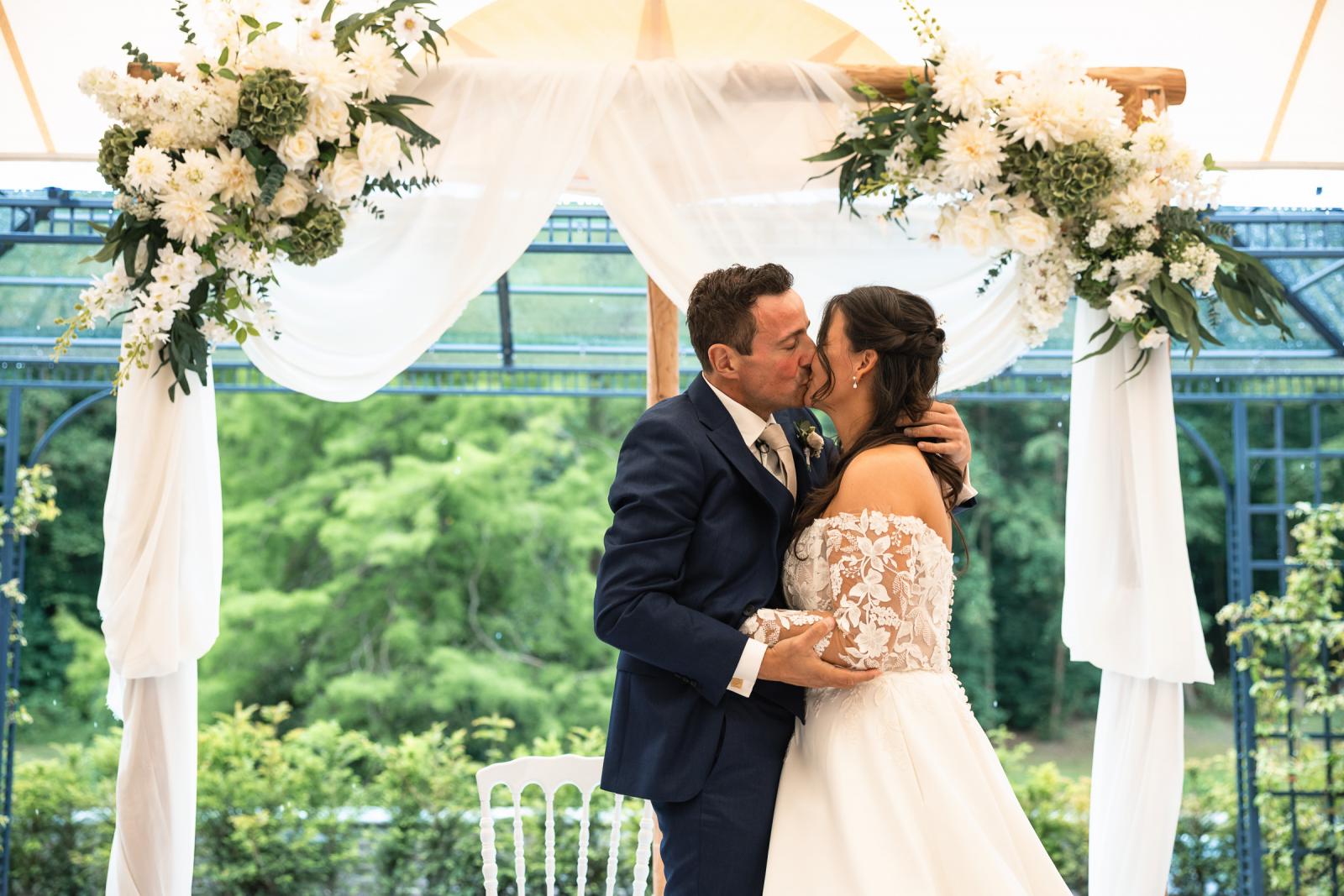 De eerste kus van George en Iris als getrouwd stel onder de prachtig met witte bloemen gedecoreerde trouwboog.
