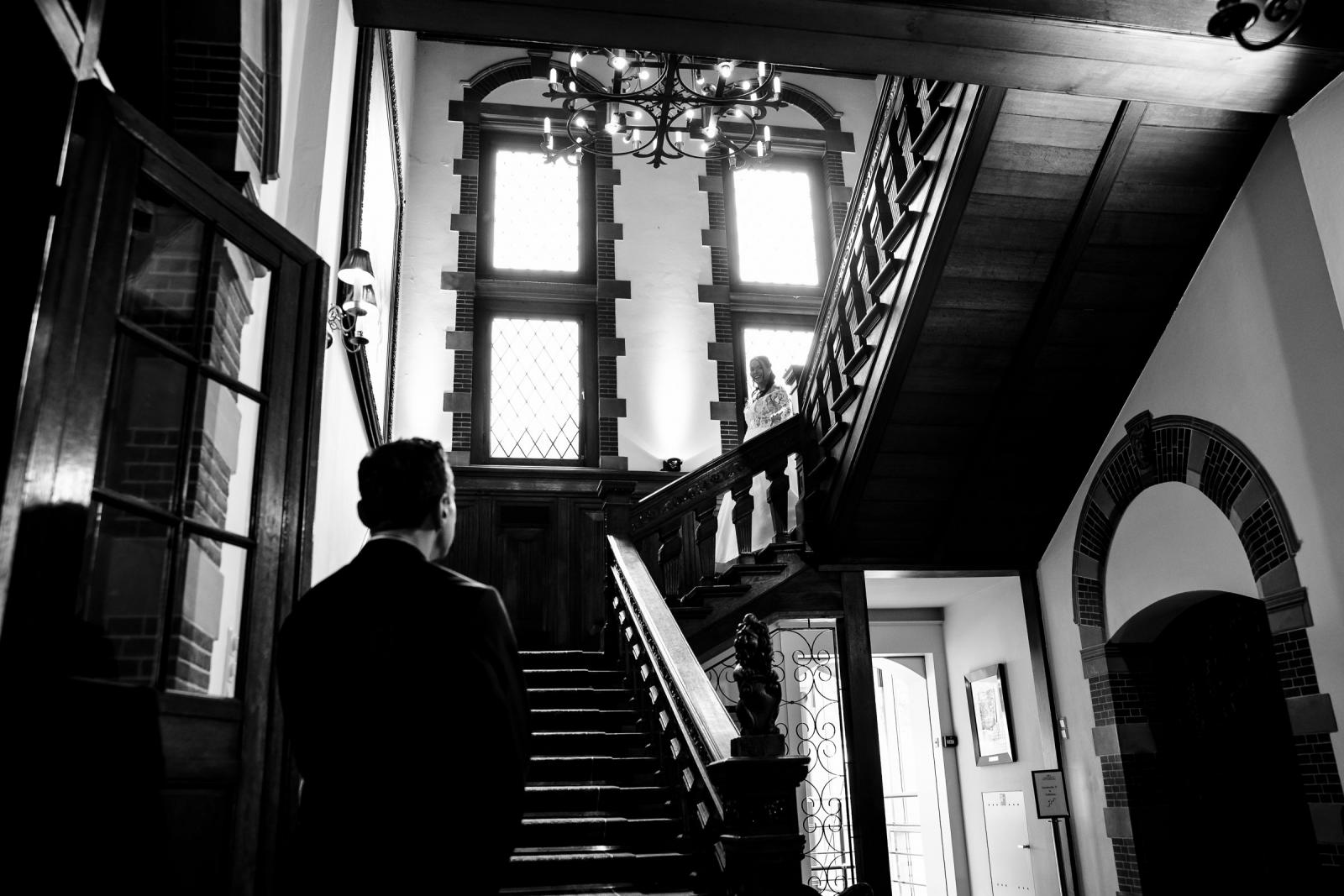 Zwart-wit foto van de first look op de houten trap in Kasteel de Wittenburg waar bruidegom George onderaan wacht op bruid Iris.