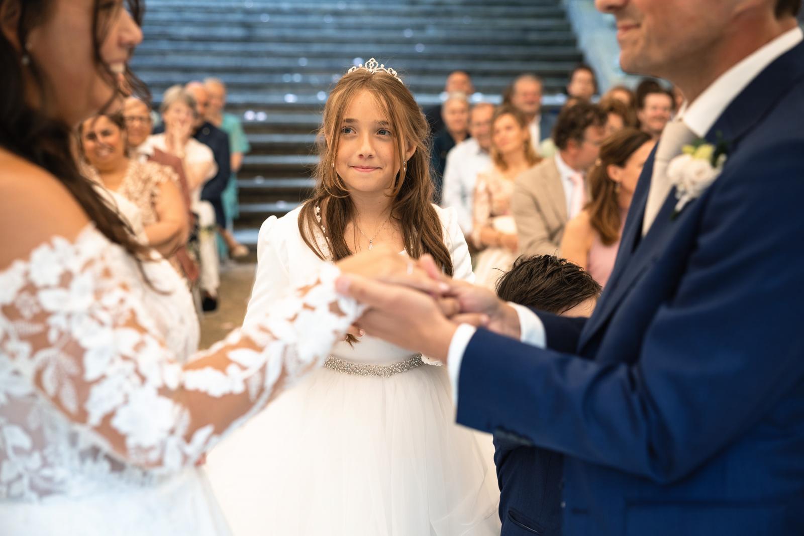 De dochter met een tiara kijkt trots toe terwijl George en Iris de trouwringen bij elkaar omdoen.