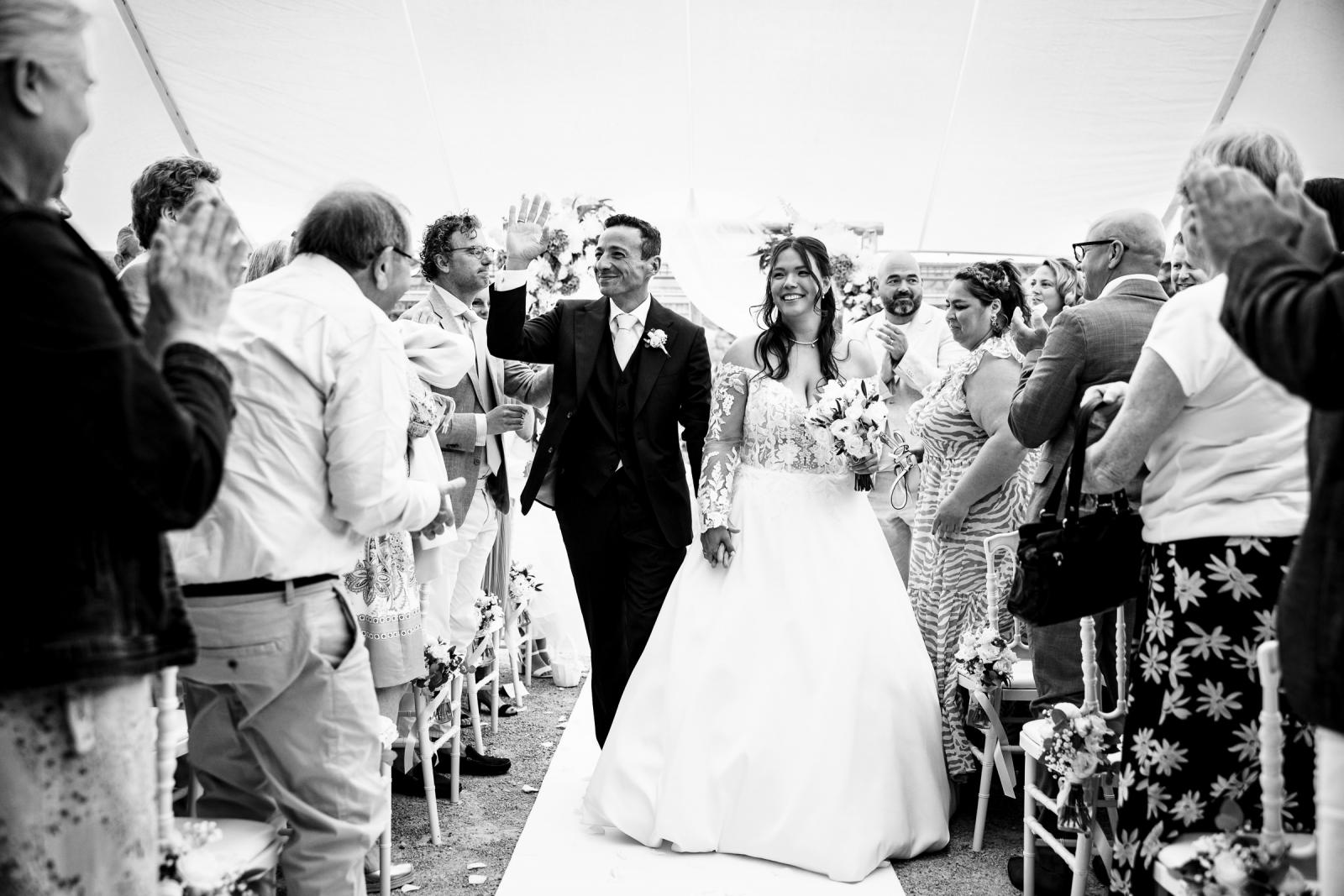 Blij zwart-wit moment waarin het kersverse echtpaar George en Iris lachend en zwaaiend over de loper loopt na de ceremonie.