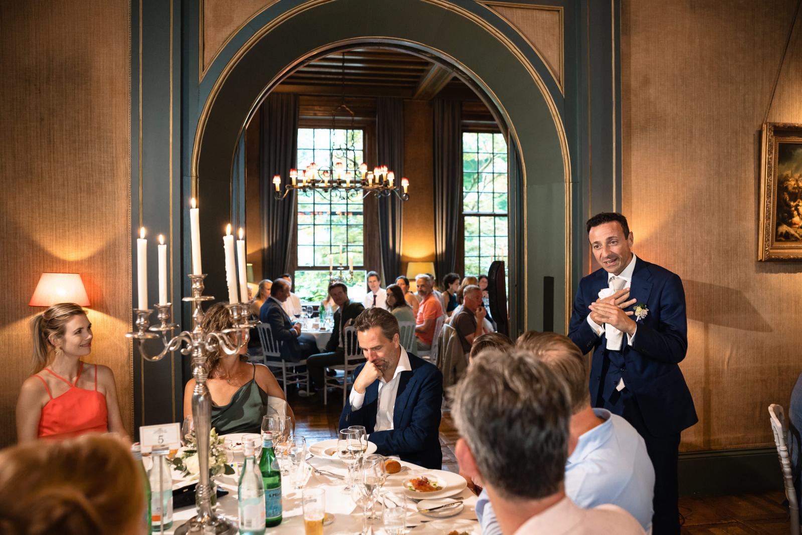 Bruidegom George spreekt met zijn hand op zijn hart de gasten toe tijdens het diner in de klassieke zaal van het kasteel.