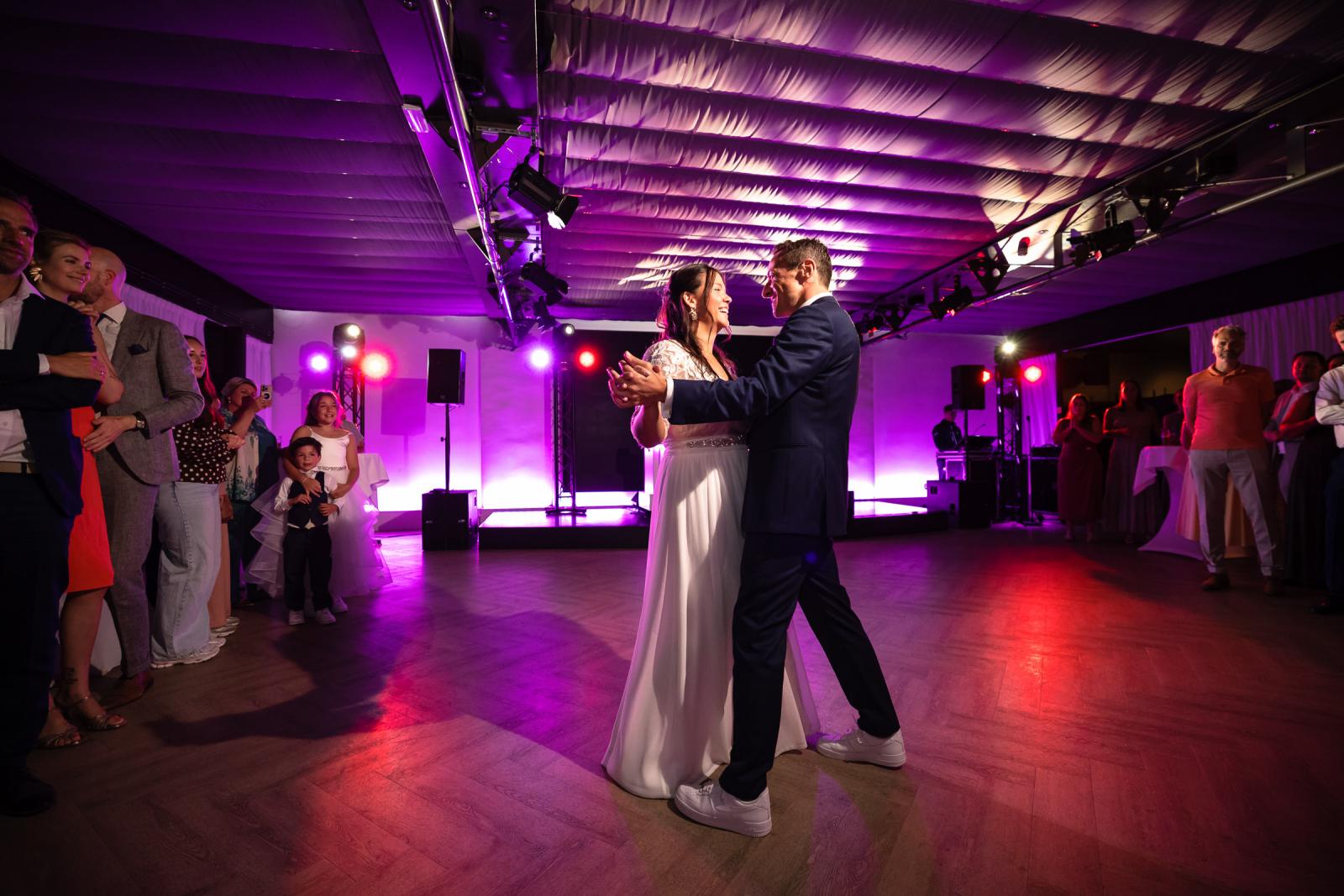 George en Iris dansen hun openingsdans in het paarse feestlicht op de dansvloer terwijl familie en vrienden toekijken.