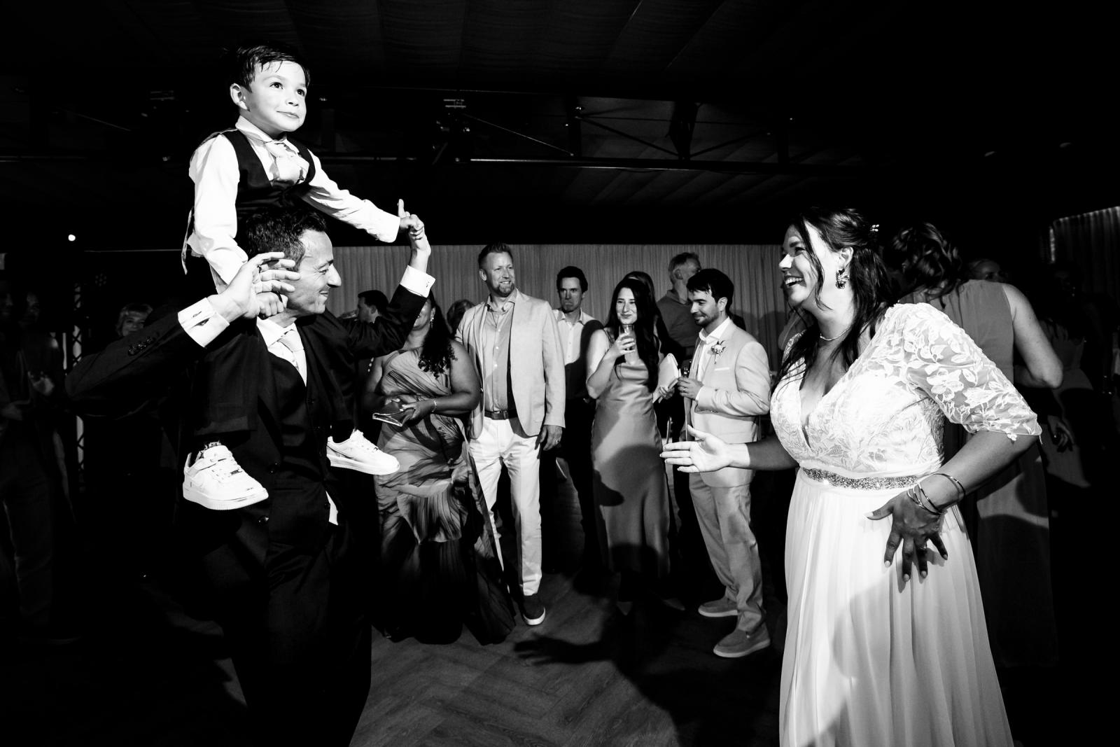 Zwart-wit foto van George die zijn zoontje op de schouders draagt en danst met Iris te midden van de feestende gasten.