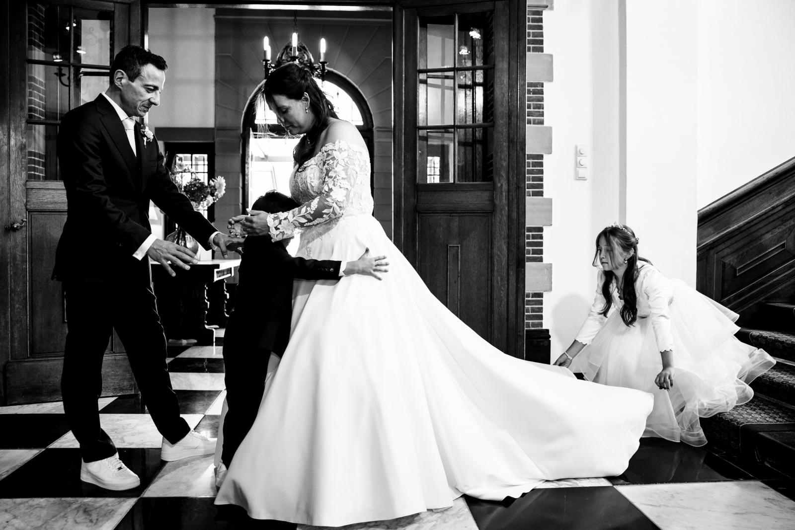Emotioneel zwart-wit gezinsmoment in de hal van het kasteel waar bruid Iris haar zoontje knuffelt en de dochter helpt met de jurk.