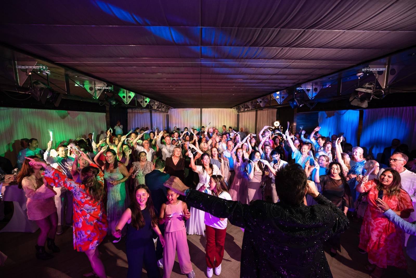 Overzichtsfoto vanaf het podium van Gerard Joling die zingt voor een laaiend enthousiaste zaal vol dansende gasten met lichtstaafjes.