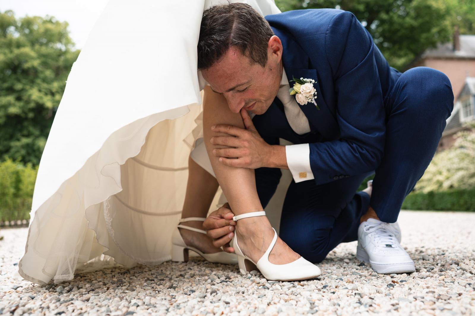 Spontane foto van bruidegom George die gehurkt onder de jurk van Iris helpt om een steentje uit haar witte bruidsschoen te halen.
