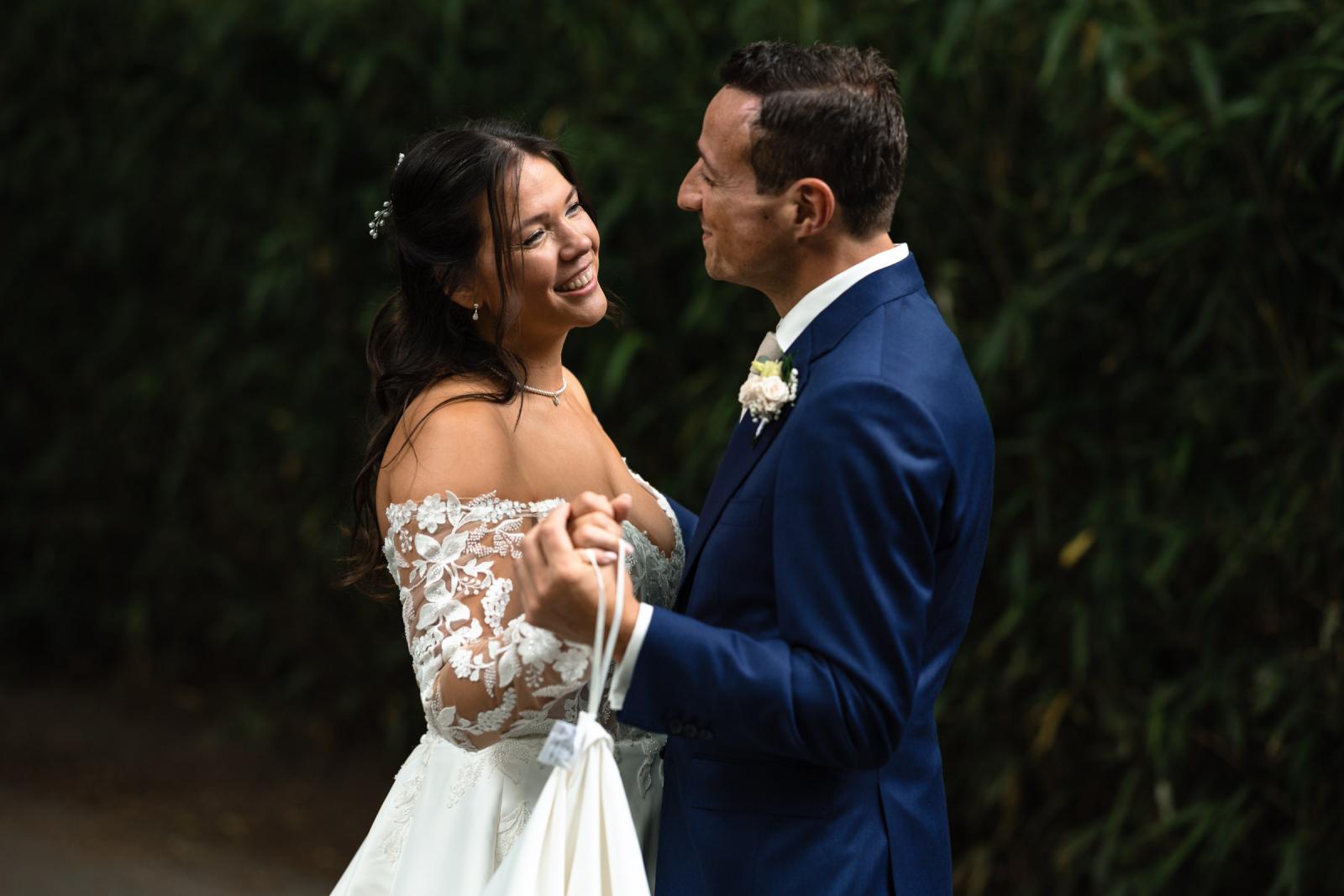 Romantisch portret van bruidpaar George en Iris die elkaar liefdevol aankijken en vasthouden voor een groene haag.