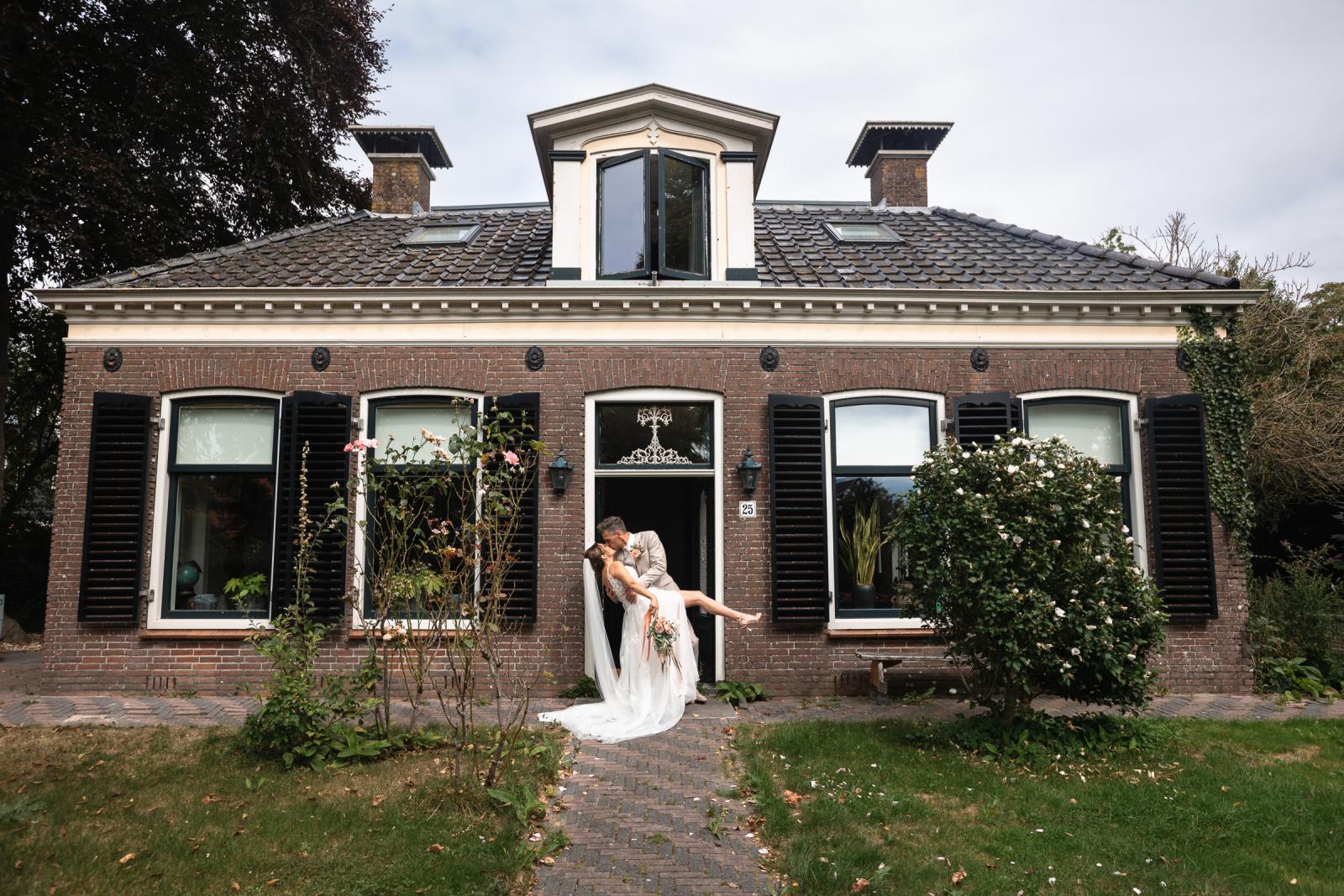 Romantische kus met dip bij de voordeur – Gerrit tilt Chantal op in haar trouwjurk voor hun huis.