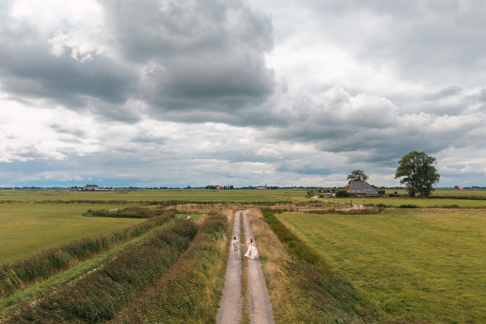 Dronefoto van Gerrit en Chantal die hand in hand over een landweg in Friesland lopen met dramatische wolken boven hen.