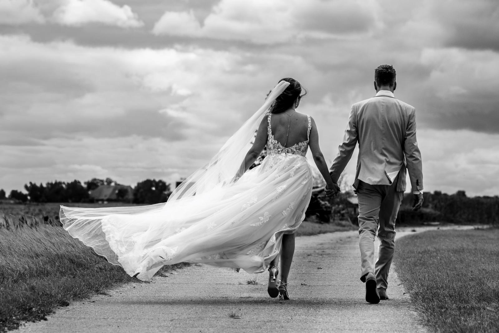 Zwart-wit foto van het bruidspaar dat hand in hand wandelt, terwijl Chantal’s sluier en jurk prachtig waaien in de wind.