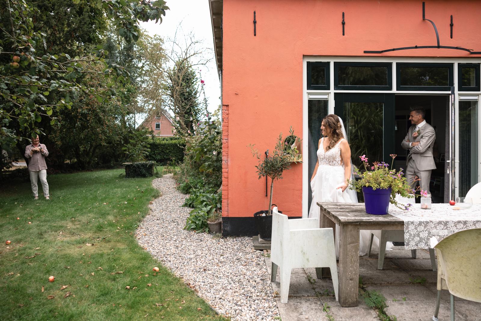 Bruid en bruidegom staan bij een oranje huis, terwijl in de tuin staat te wachten op zijn dochter.