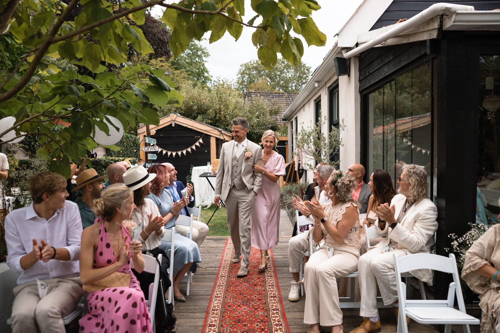 Bruidegom loopt trots de ceremonie binnen aan de arm van zus, toegejuicht door gasten.