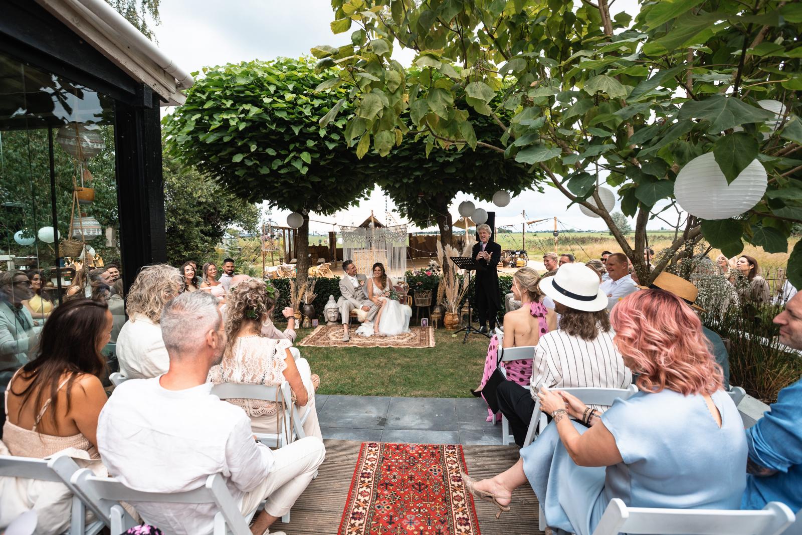 Overzicht van de trouwceremonie in de tuin met het bruidspaar voor een boog van bomen.