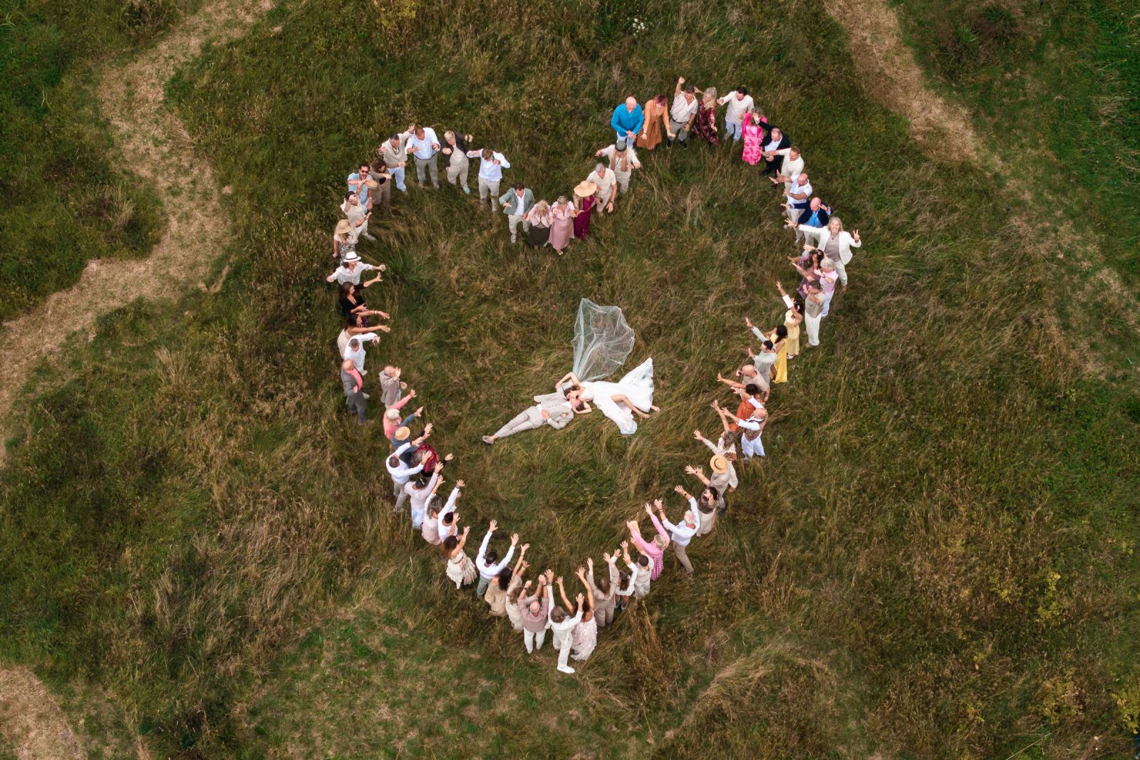 Dronefoto van bruidspaar liggend in het gras, omringd door gasten die een hartvorm maken.