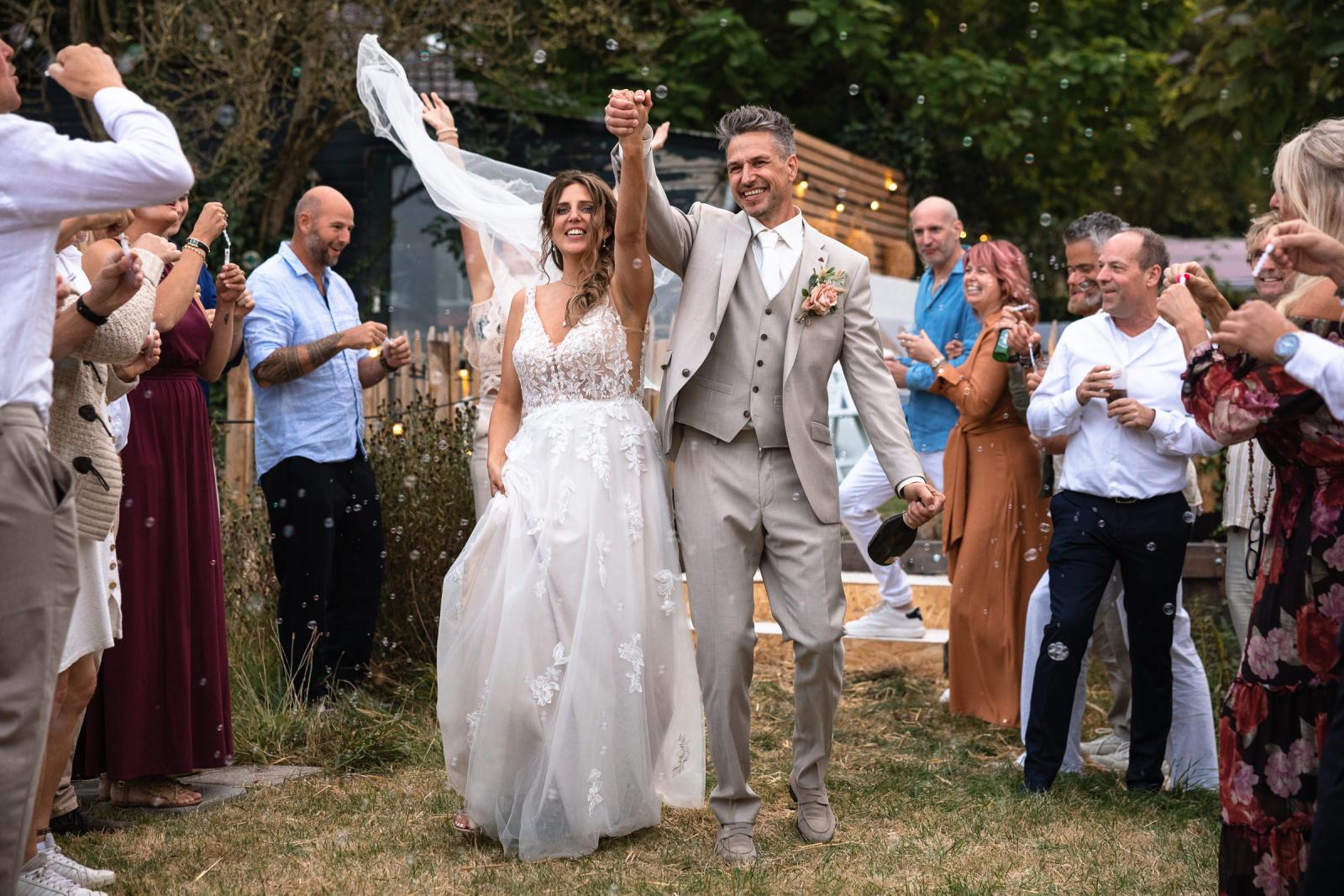 Bruid en bruidegom lopen juichend hand in hand door een haag van gasten met bellenblaas.